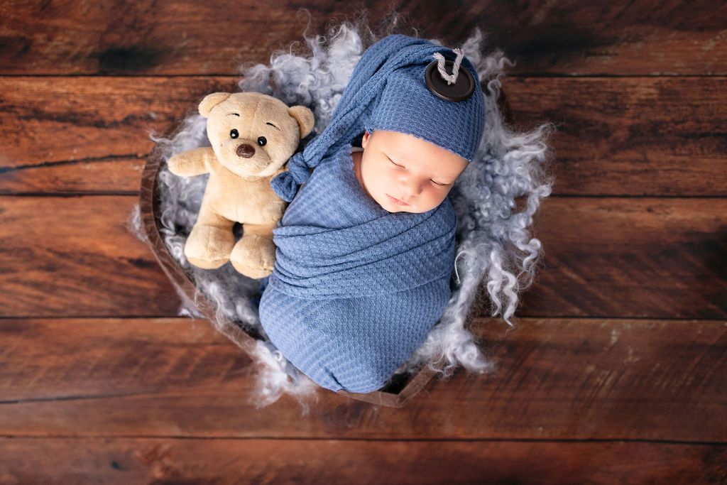 Newborn swaddled in blue lies on rustic wooden floor with soft grey fluff decoration.