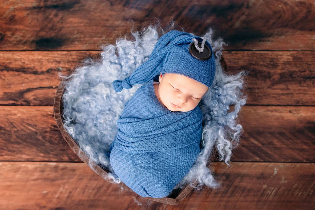 Overhead view of baby in blue wrap nestled in grey fluff on dark wooden flooring.