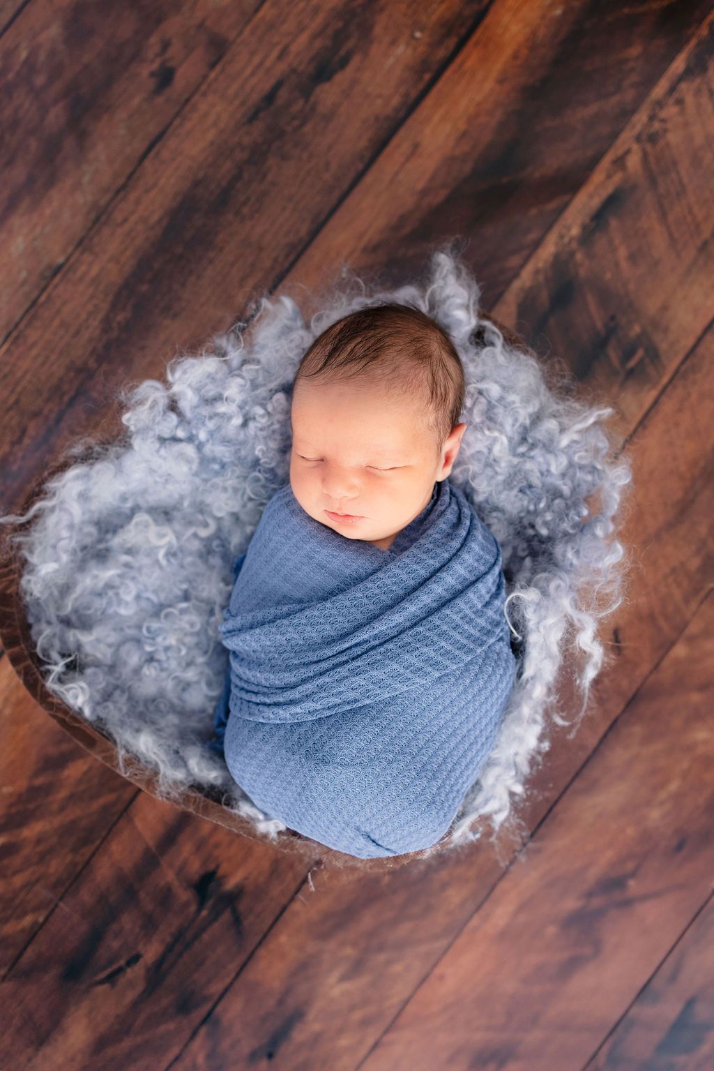 Bird's eye perspective of newborn in blue wrap surrounded by fluffy grey material on wood floor.