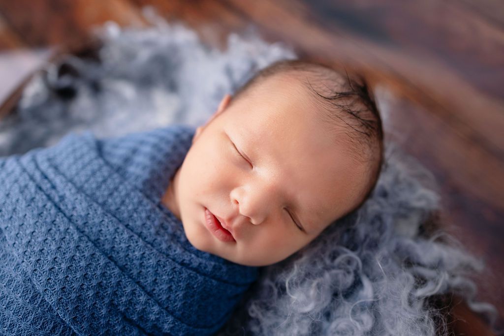 Sleeping baby wrapped in navy blue textile on rustic wooden surface with grey fluff accents.