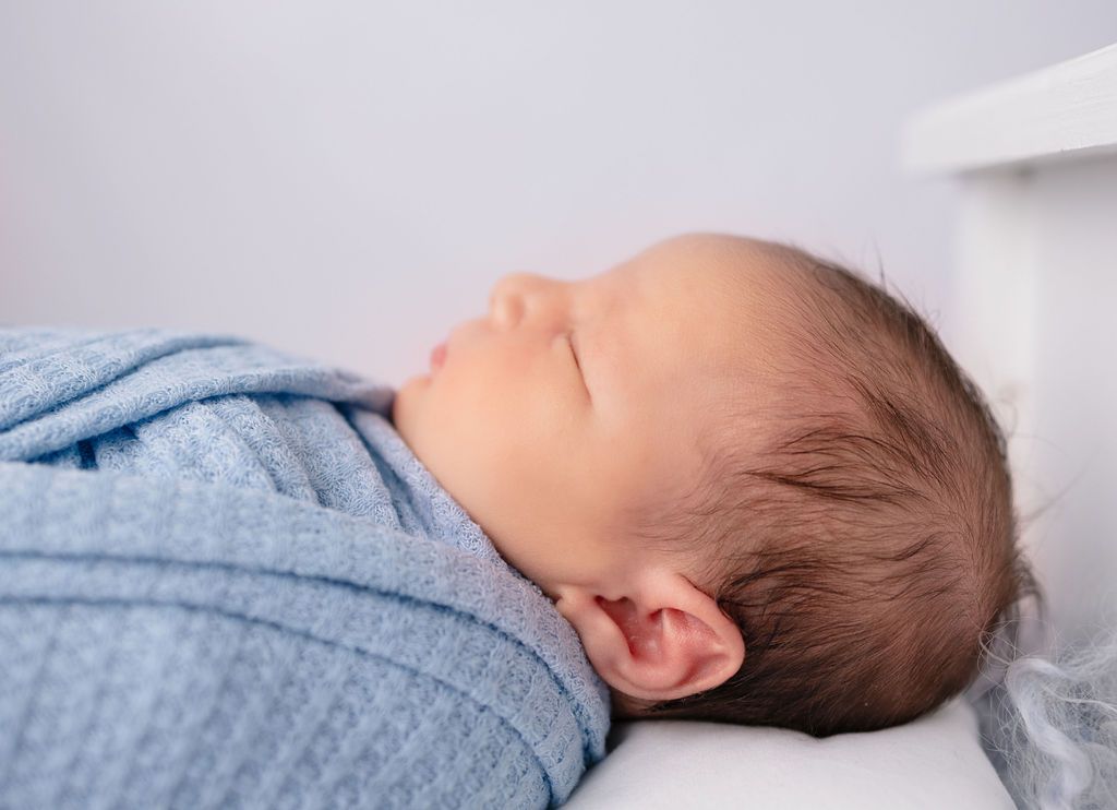 Sleeping baby wrapped in light blue swaddle on white wooden bed.