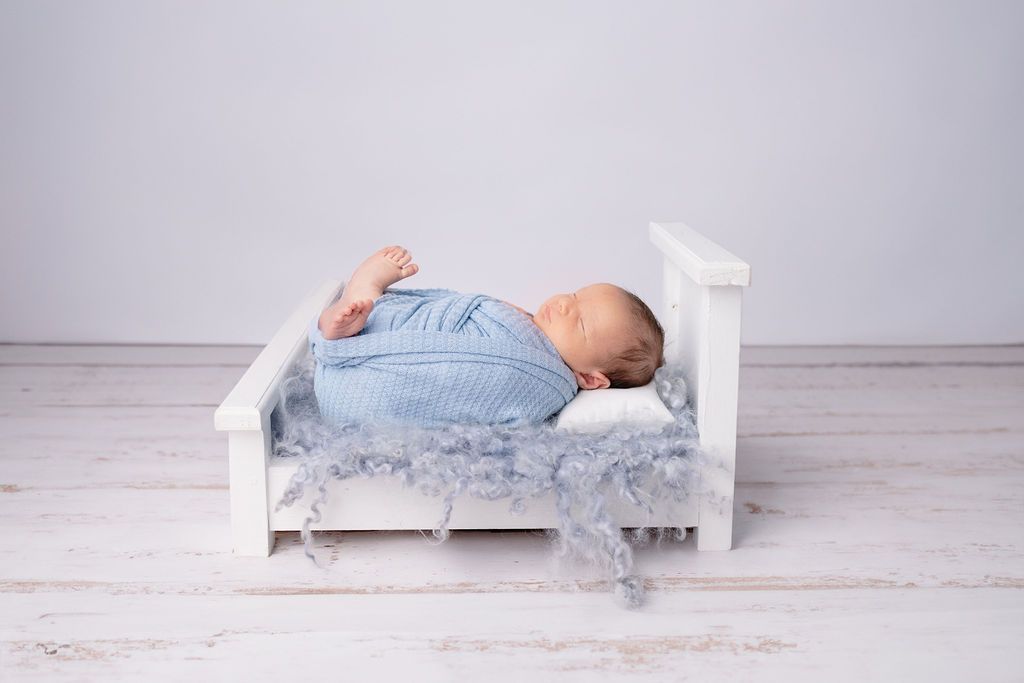 Small white wooden bed with sleeping infant in blue wrap against light gray background.