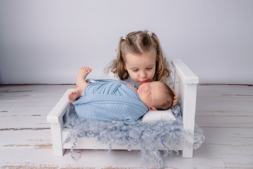 Young child peeking over edge of white wooden bed at sleeping baby.