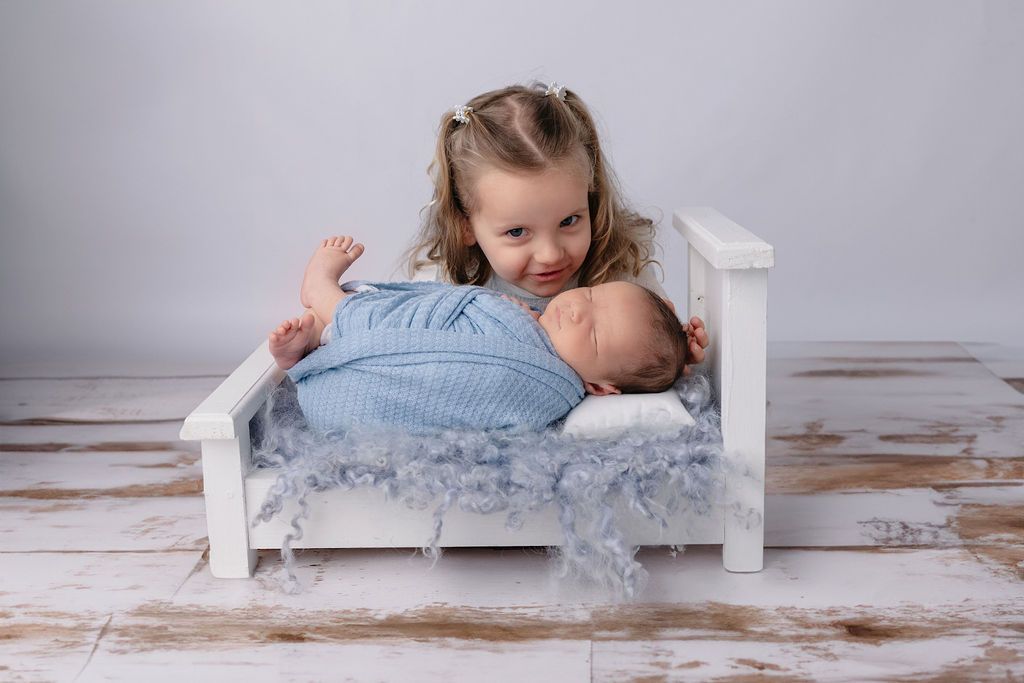 Child curiously looking over edge of white bed at sleeping infant.