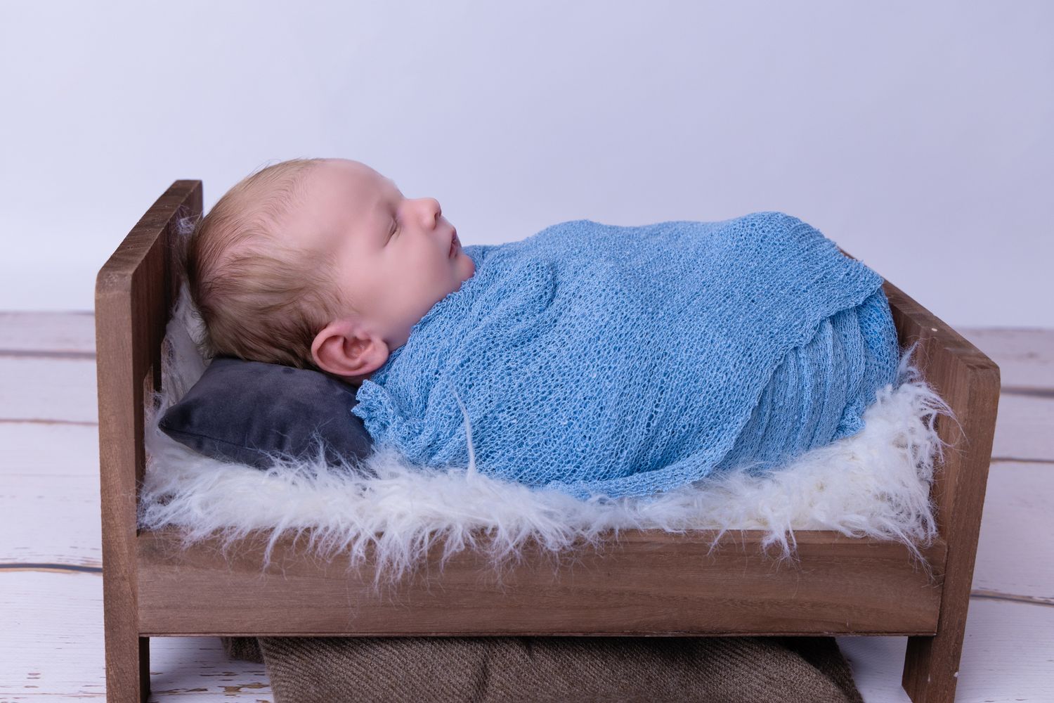 Peaceful baby sleeping on wooden platform with blue wrap against light background.