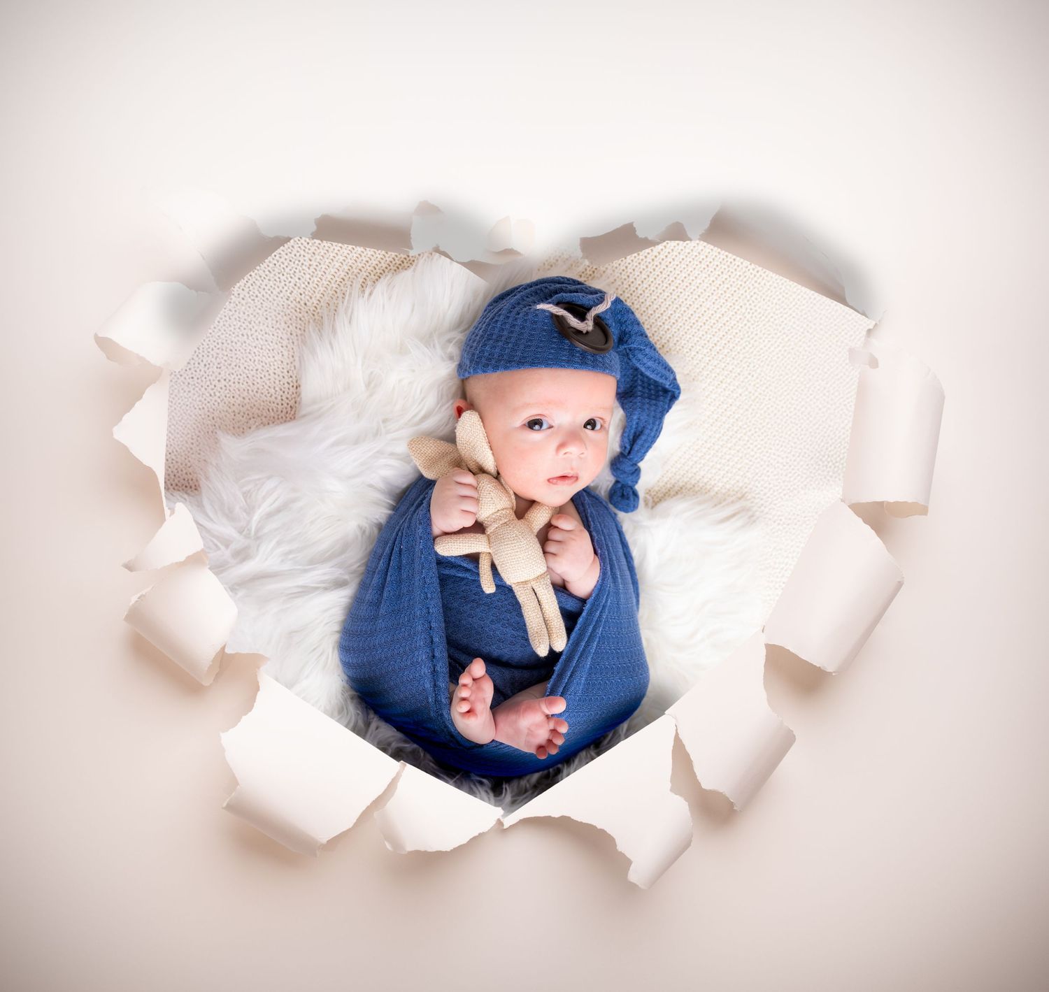 A baby in blue overalls peeks through torn white paper creating a creative photo backdrop effect.