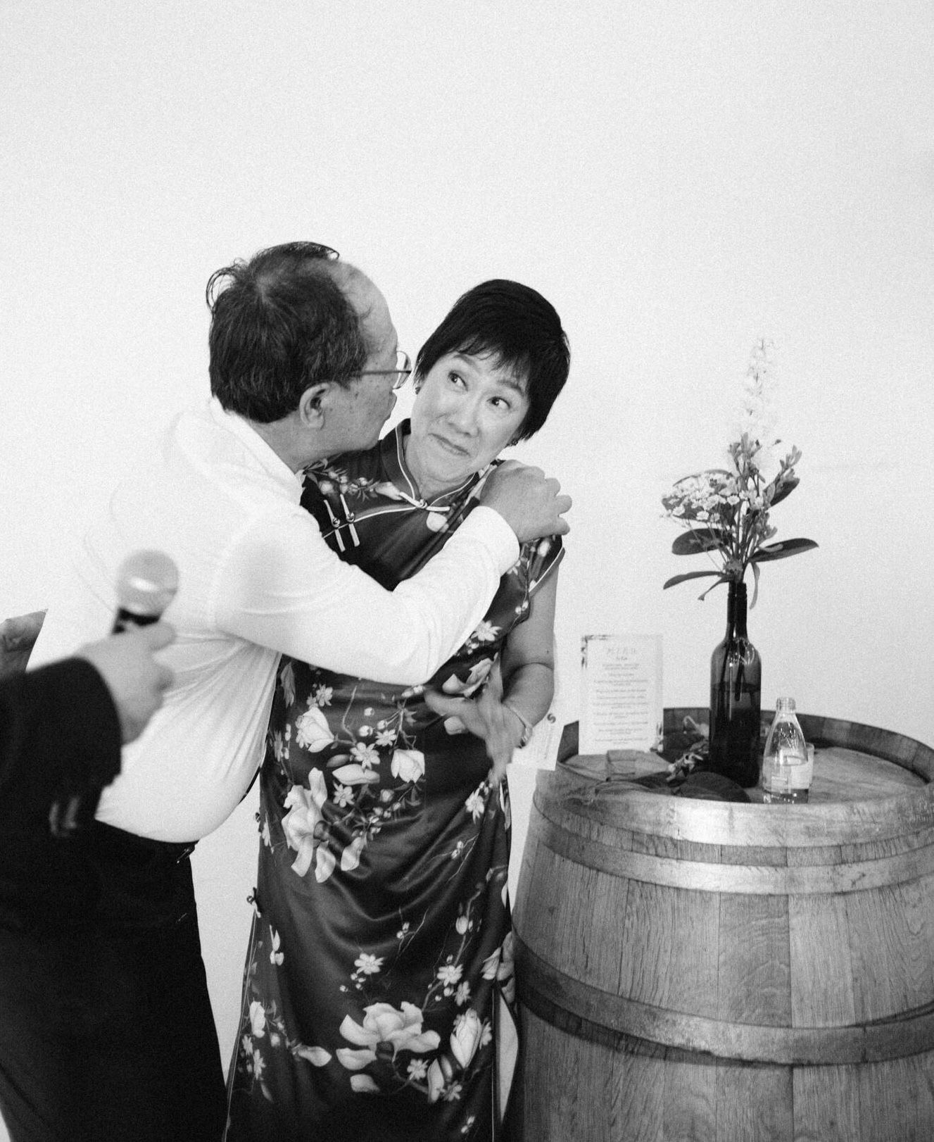 Cheeky photo of bride's dad kissing on his wife's cheek. Heartwarming black and white photo at the indoor wedding venue in melbourne's north