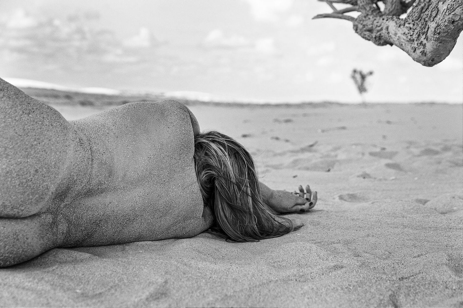 Black and white artistic photograph of a person lying on a sandy beach with distant horizon visible in background.