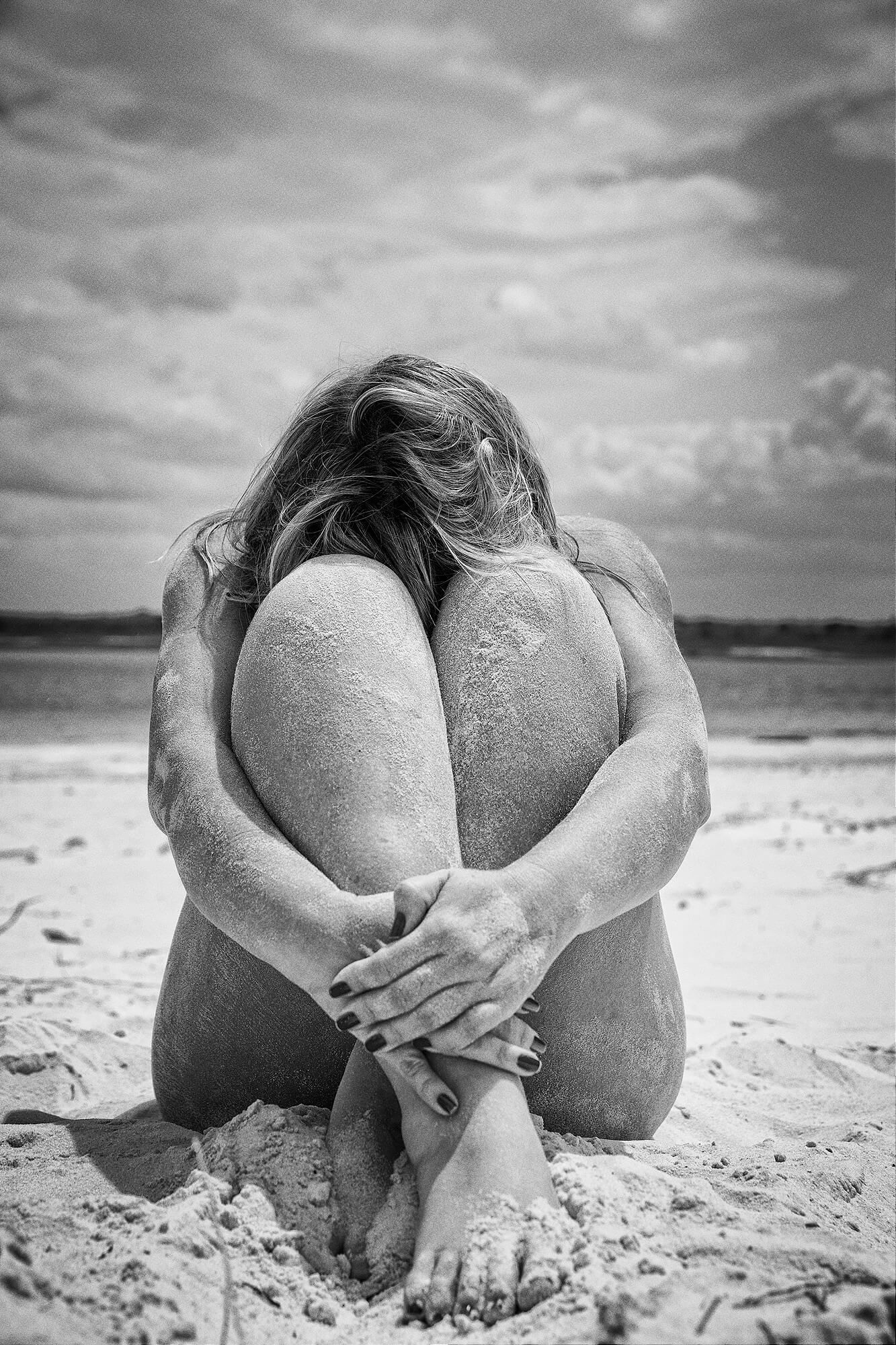 Black and white photo of someone curled up on a sandy beach under dramatic cloudy skies.