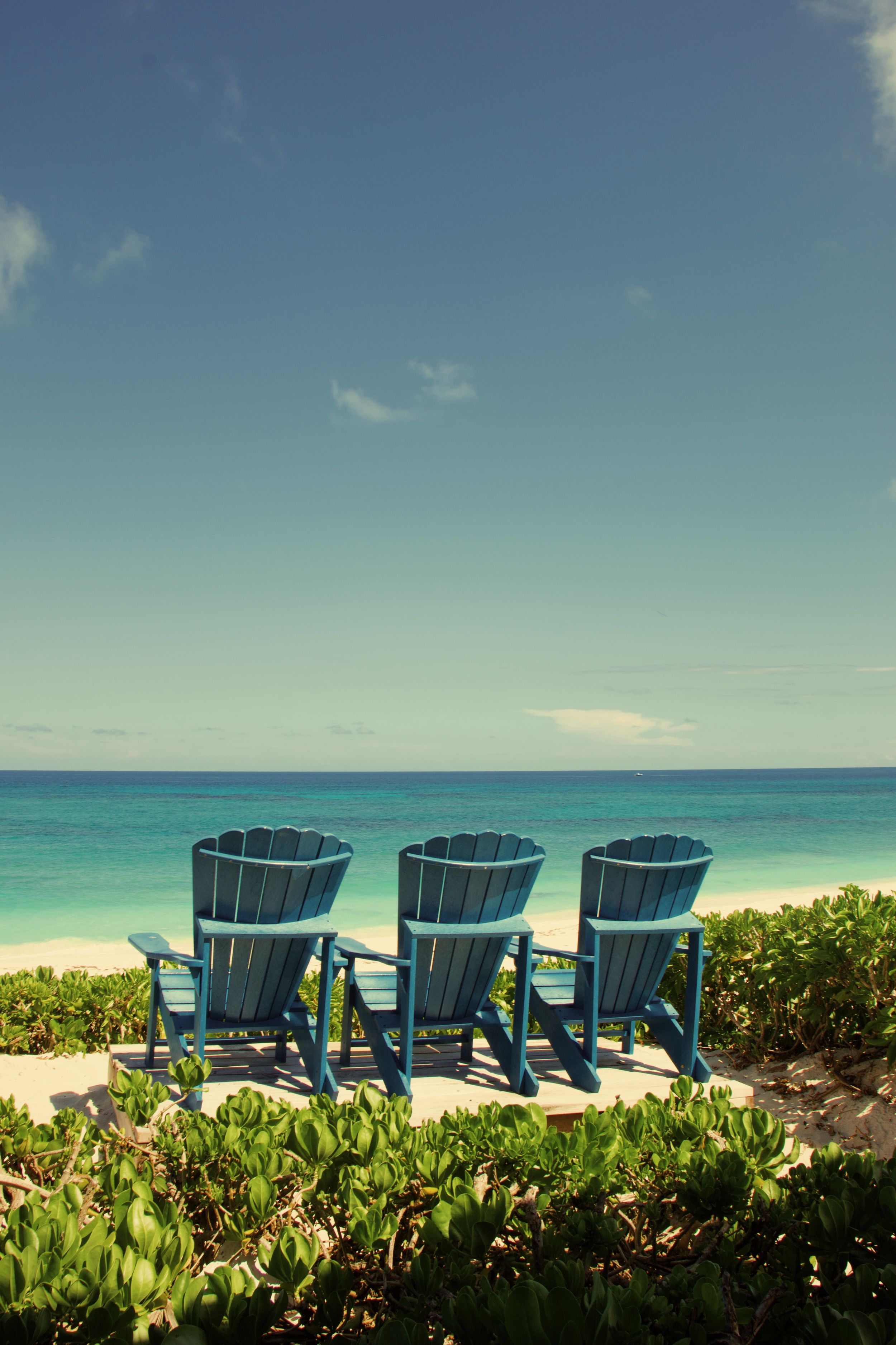 Three blue Adirondack chairs overlook a pristine turquoise ocean beach with tropical foliage in the foreground.