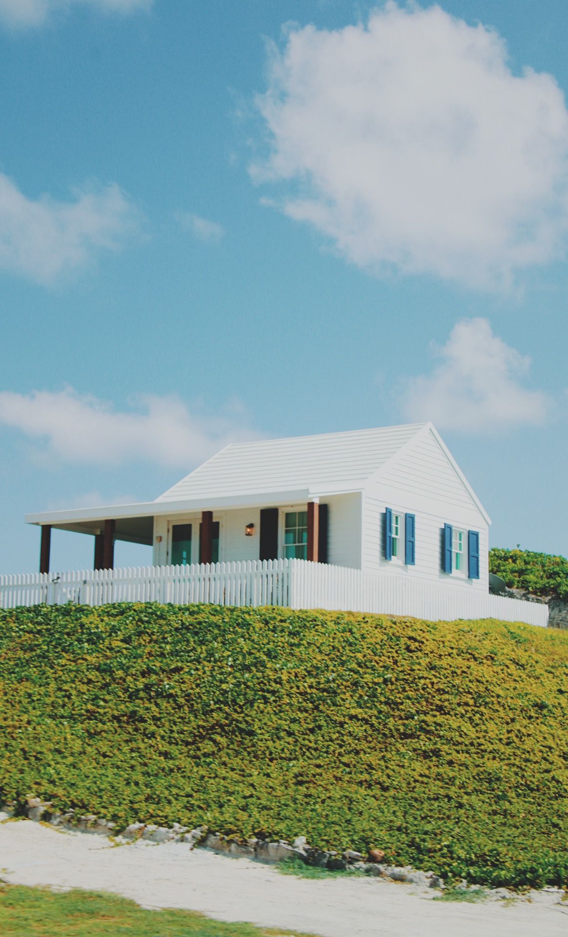 White cottage with blue shutters sits atop grassy hill against bright blue sky.