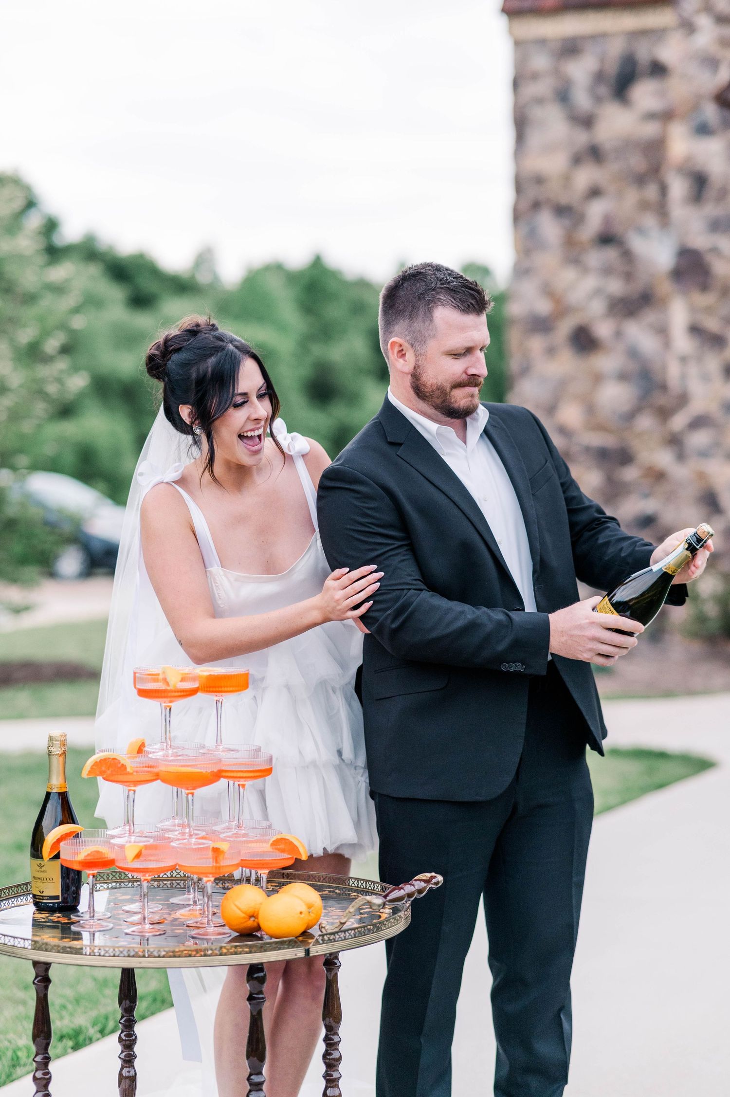 Newlyweds celebrating with champagne and a tiered orange dessert display at an outdoor wedding reception.
