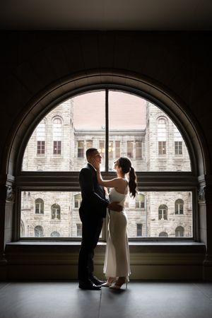 Silhouetted couple embraces in front of arched window overlooking historic stone building architecture.