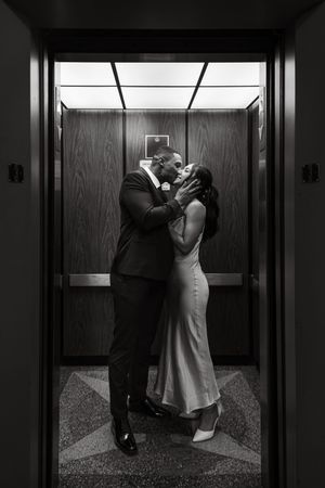 A couple shares a romantic kiss inside an elevator with wood paneled walls, captured in black and white.