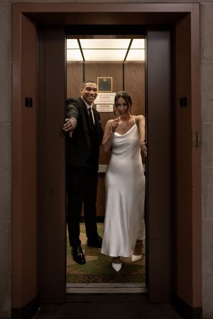 A couple in wedding attire poses in a brightly lit elevator, with the bride in white and groom in black suit.