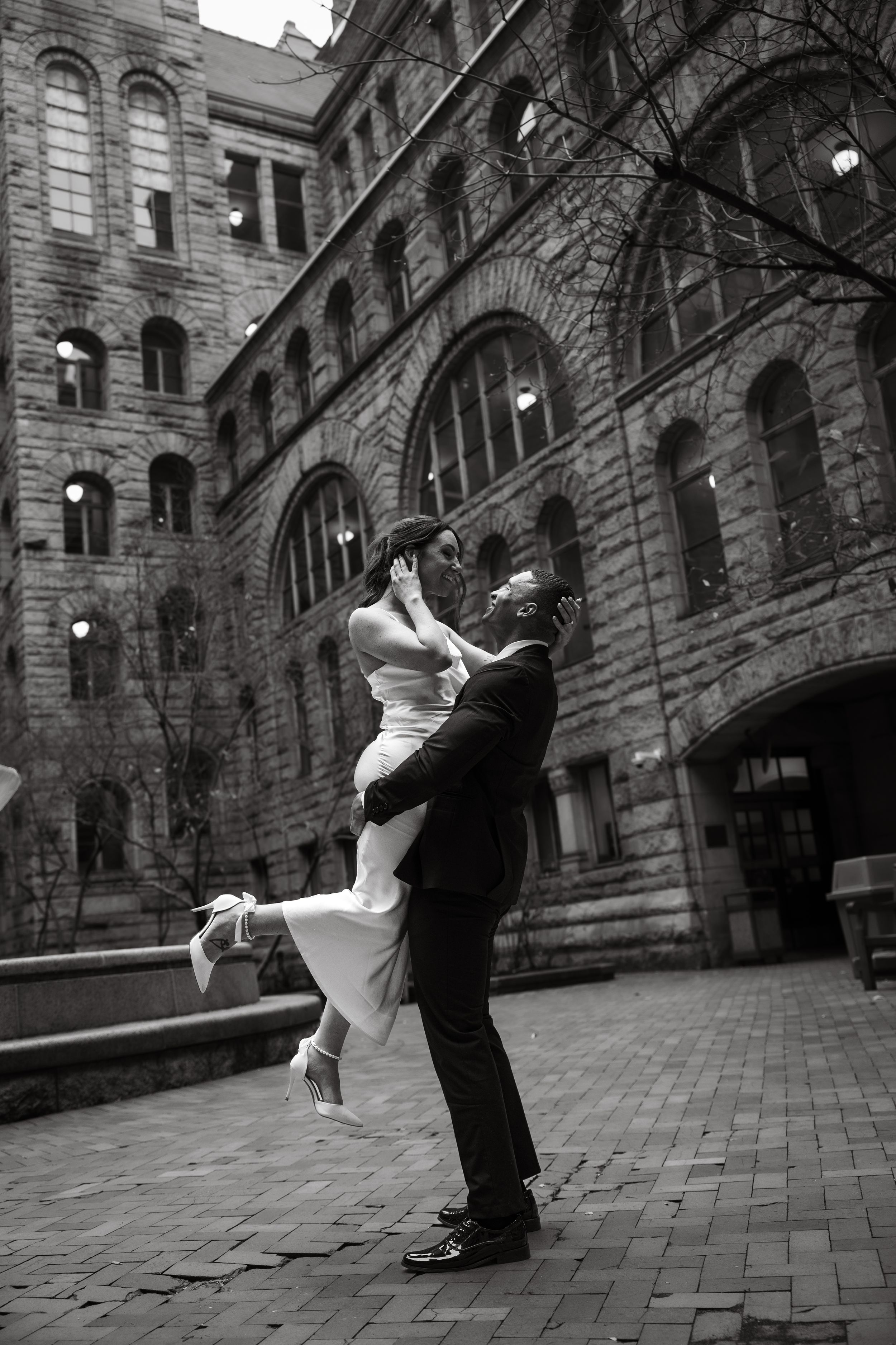 A joyful wedding couple dancing and twirling outside a historic stone building with arched architecture.