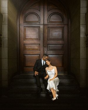 Two people in formal attire sit together on stone steps in front of a grand wooden double door in moody lighting.