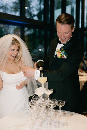 A couple at their wedding reception pour champagne into a tower of crystal glasses at their celebration.
