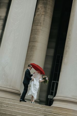 A couple shares an intimate moment under a red umbrella between classic white stone columns.