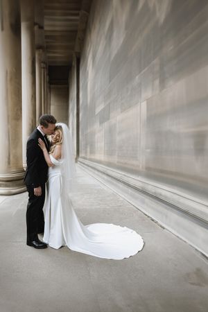 Couple in wedding attire embrace in elegant colonnade hallway with flowing white dress train against stone architecture.