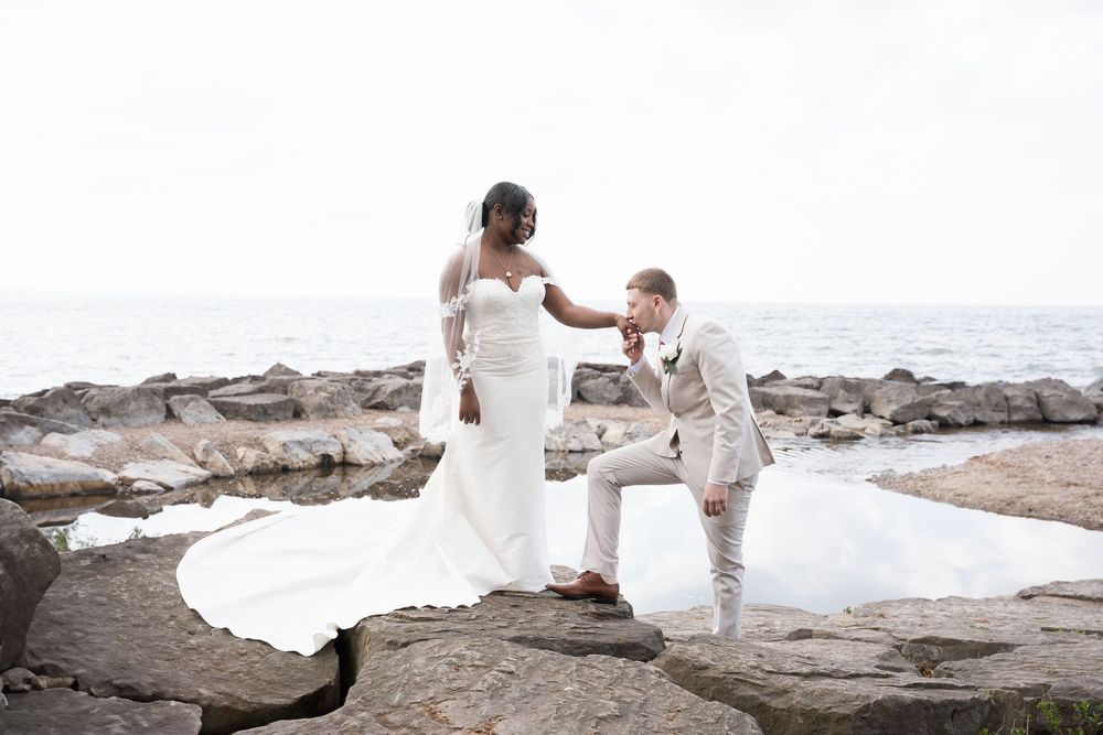 Romantic beach wedding photoshoot with couple posing on rocks near ocean at sunset in white dress and beige suit.