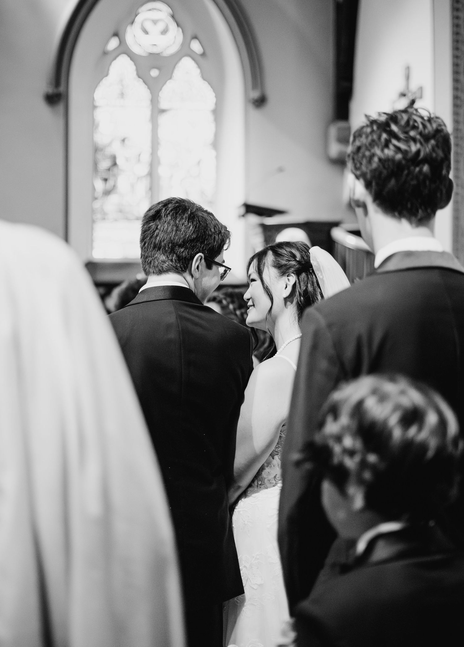 Black and white photo of couple during wedding ceremony in church with arched stained glass window in background.