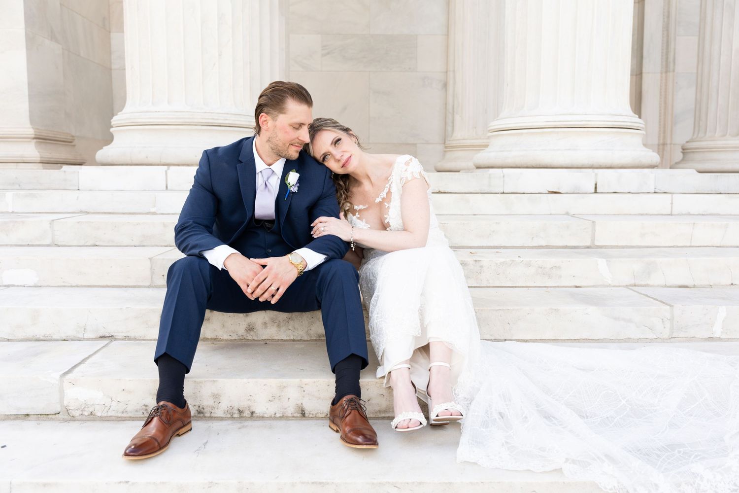 A couple in wedding attire sits together on white stone steps outside a classical building.