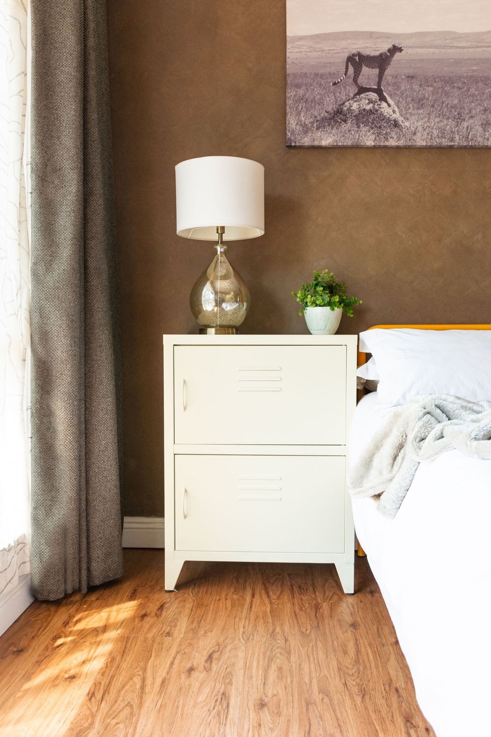 White metal locker-style nightstand with gold lamp beside a bed, brown textured wall, cheetah canvas art, hardwood floor.
