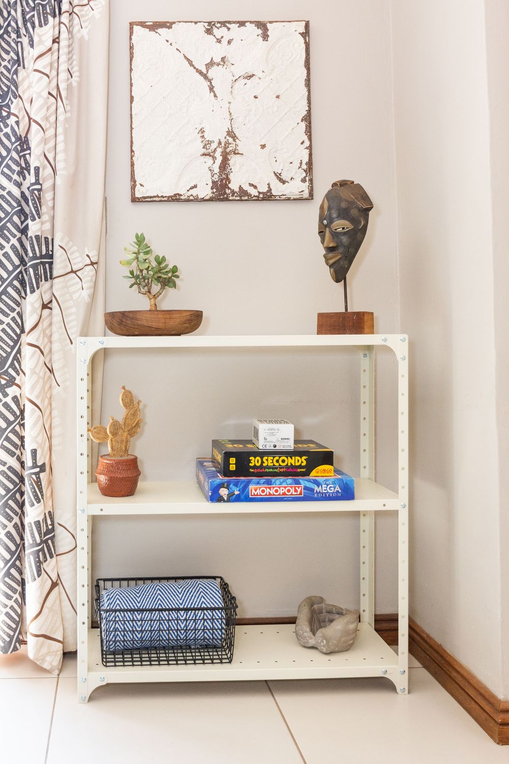 White metal shelf with Monopoly board game, small plants, woven basket, and African mask decor against beige wall.