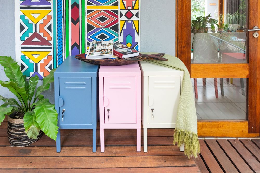 Three colorful metal lockers in blue, pink, and cream on a wooden porch with tropical plants and vibrant African art.