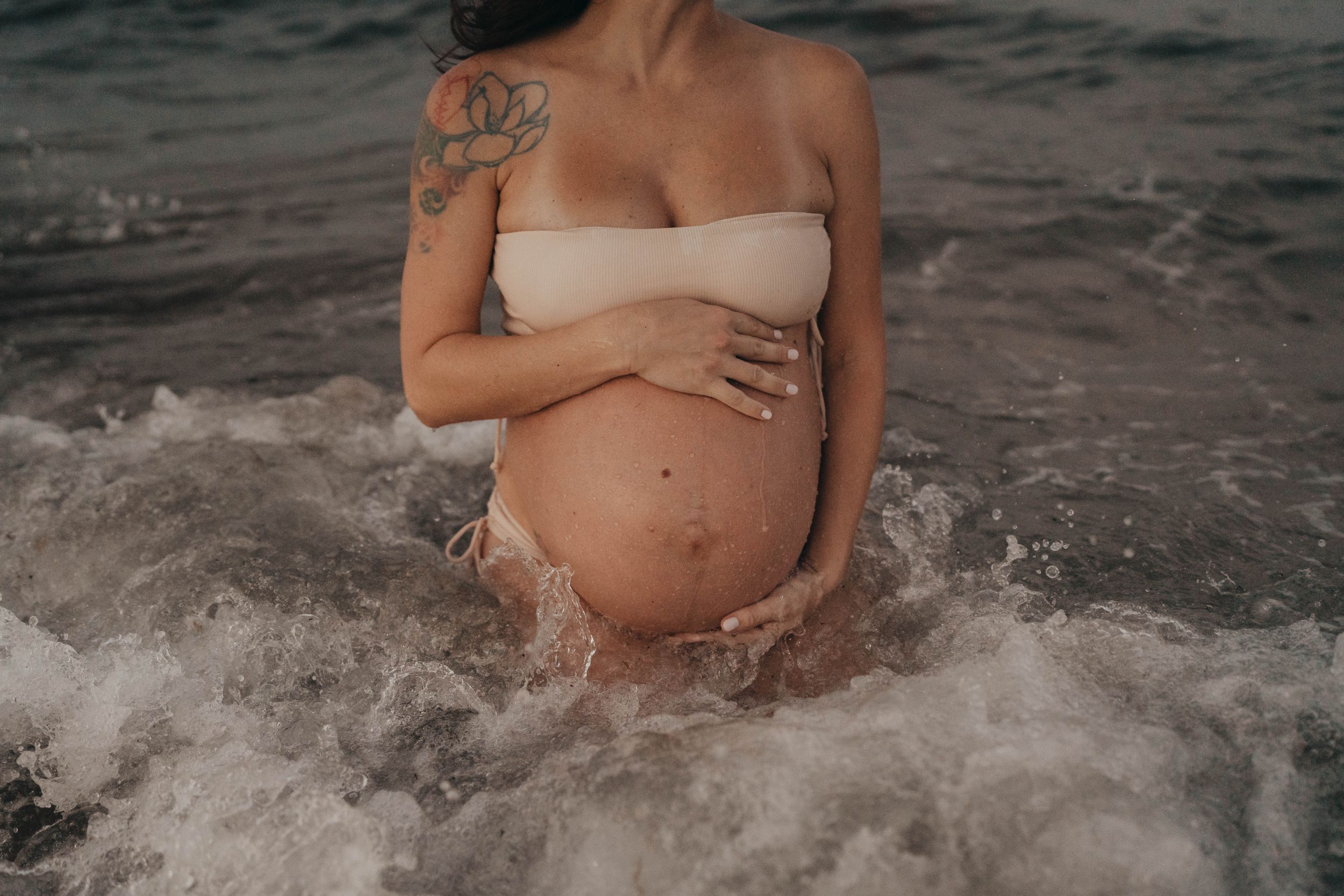 A pregnant woman stands in ocean waves wearing a nude bandeau top.