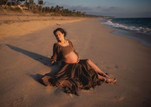 Expectant mother in flowing dress relaxing on a scenic beach at sunset.