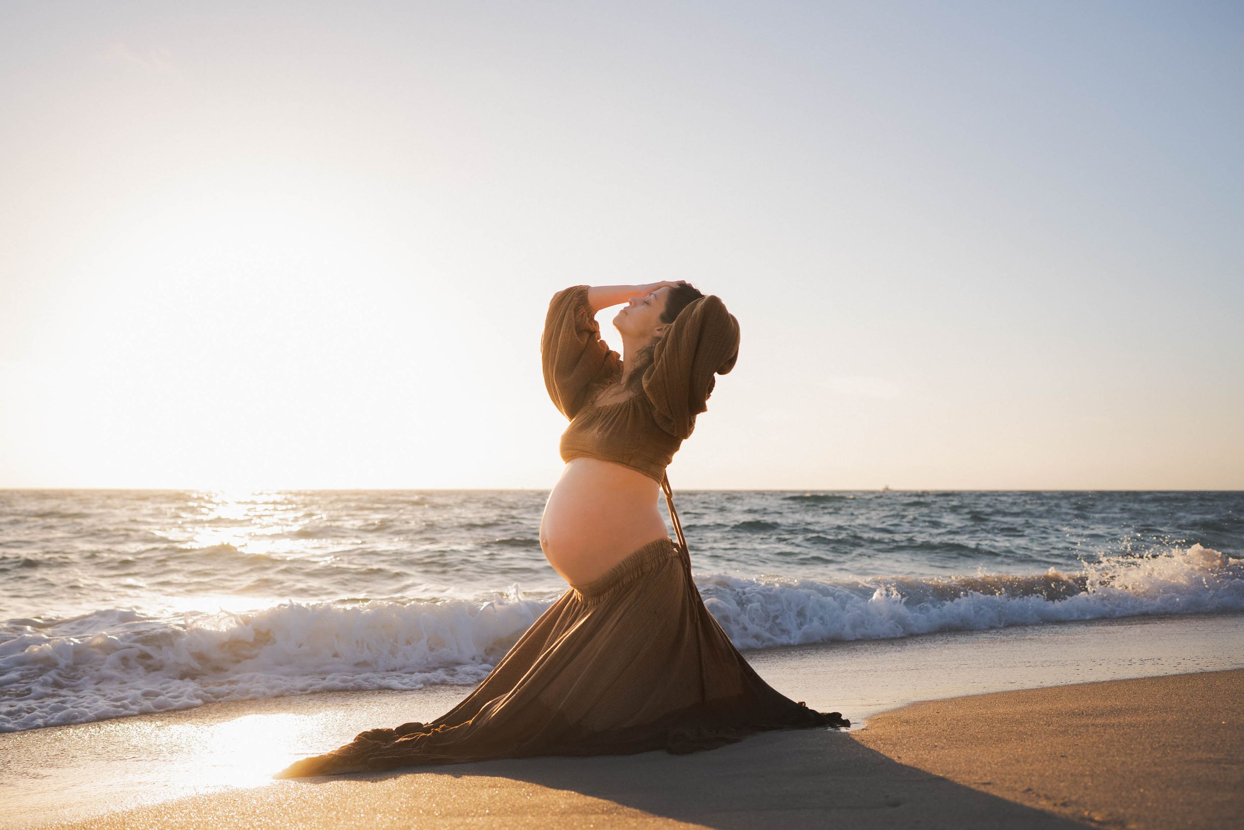 Silhouette of pregnant figure against ocean sunset, waves crashing in background.