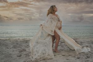 Maternity beach photo with flowing lace dress against cloudy ocean backdrop.