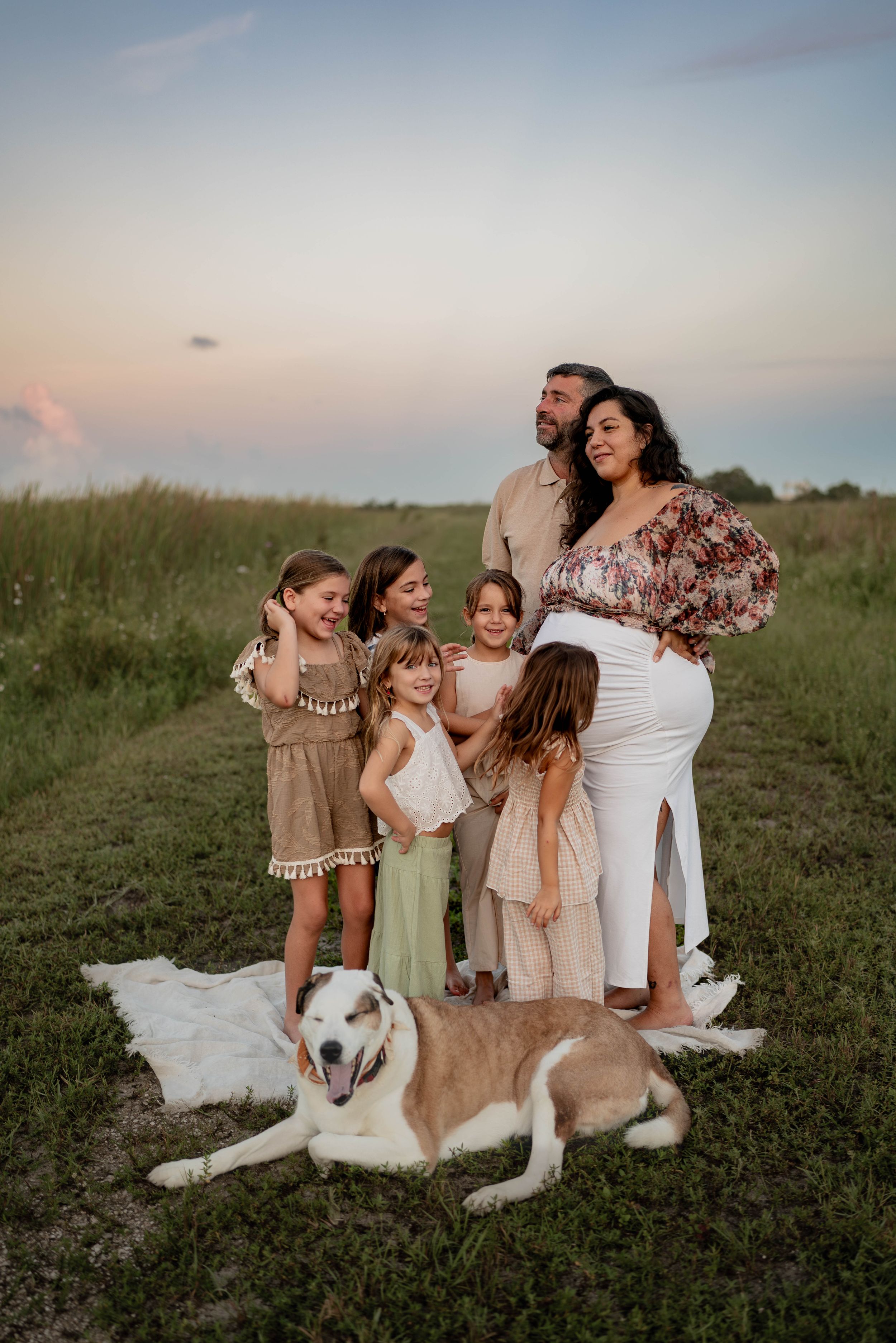A family poses together in a field at sunset with their husky dog.