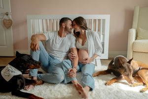 A couple sits on a white rug with dogs relaxing by a baby crib.