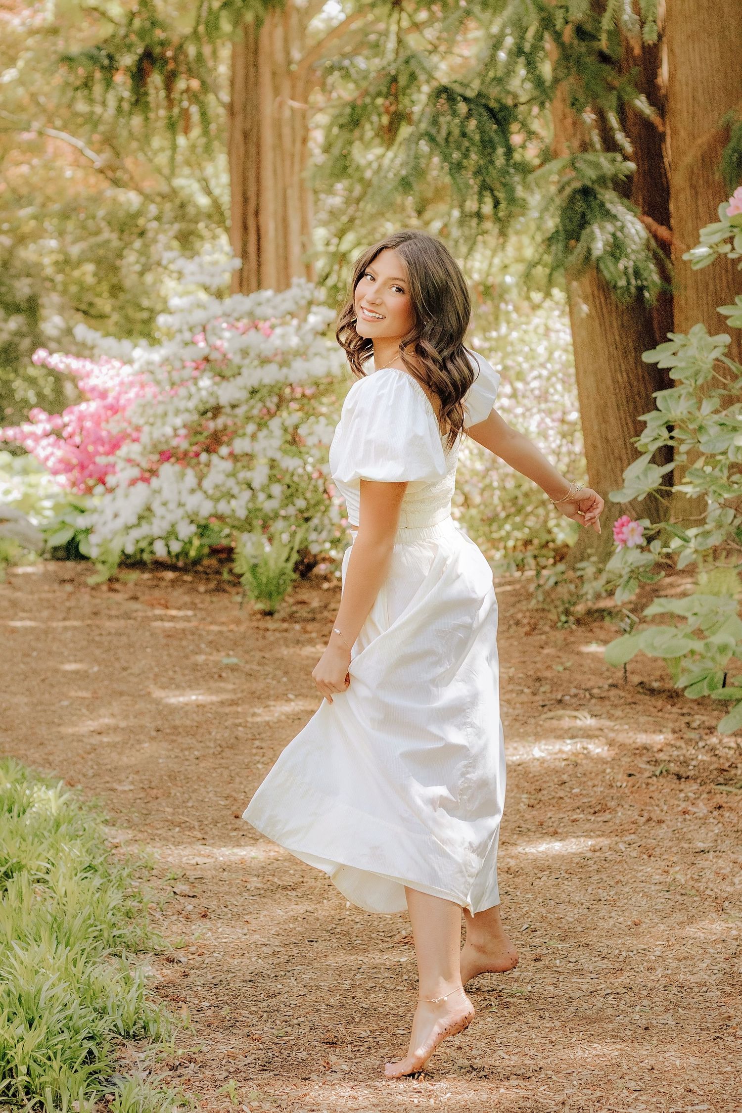A brunette woman in a white dress twirls barefoot on a garden path among blooming flowers.