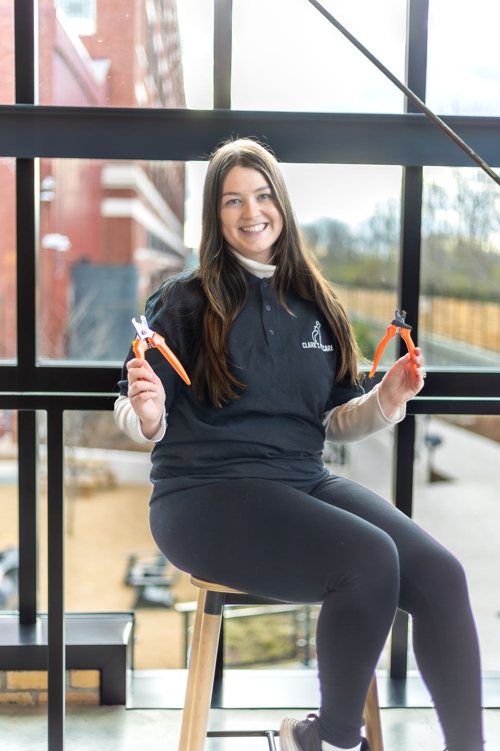 Team member in branded uniform demonstrates scissors while seated on stool near windows.
