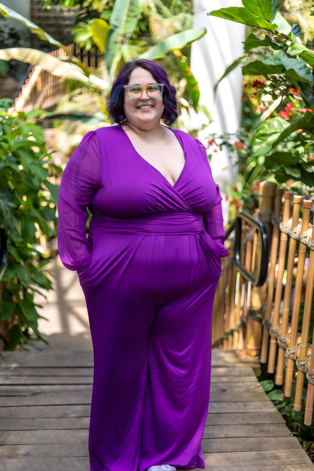 Person in purple wrap dress standing on wooden walkway surrounded by lush tropical plants.