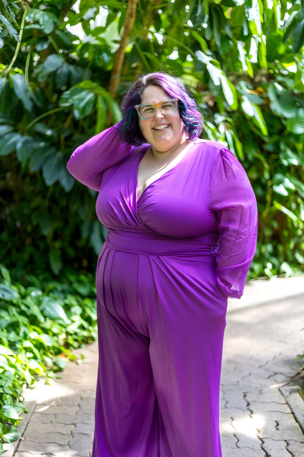Person wearing flowing purple jumpsuit posing in garden setting with tropical foliage.