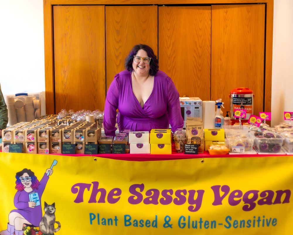 A vendor stands behind a yellow Sassy Vegan booth displaying plant-based and gluten-sensitive food products.