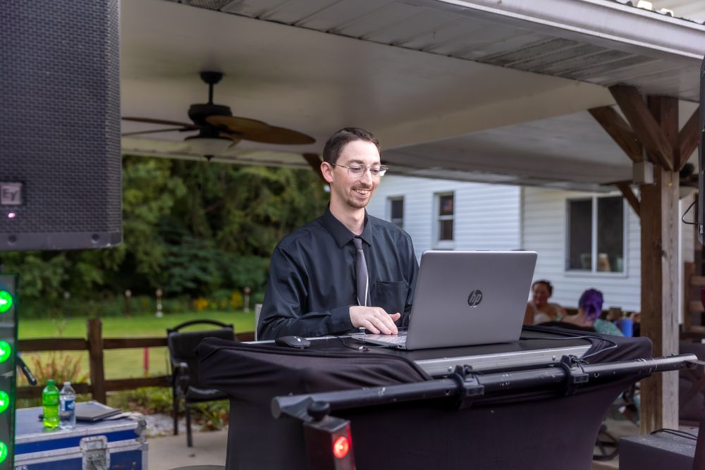 DJ performing with laptop and professional audio equipment at an outdoor covered venue setup.