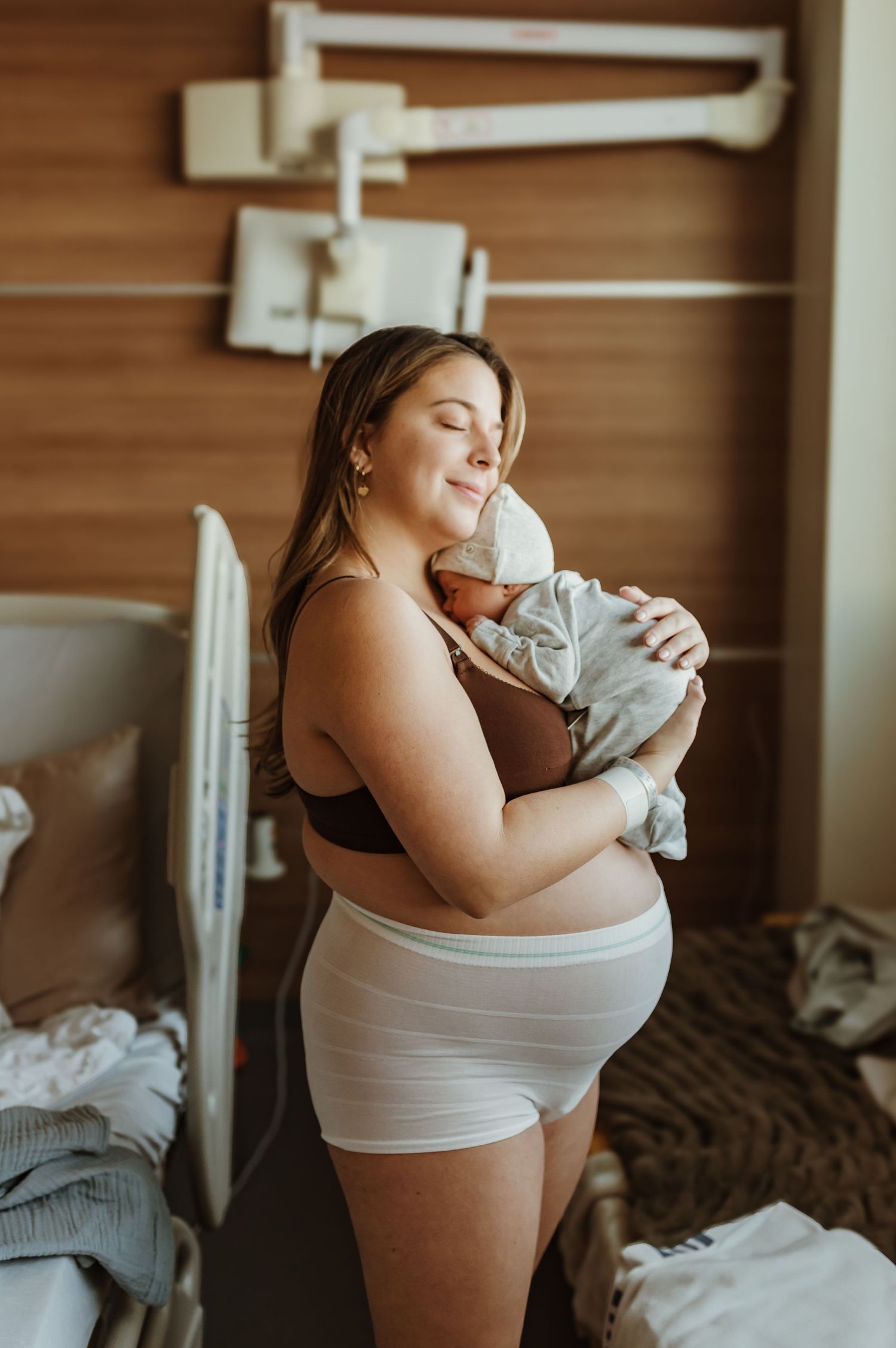 Pregnant woman in nursing bra and shorts holds newborn in hospital room.
