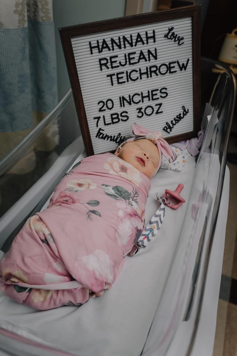 Newborn rests in hospital bassinet beneath birth statistics board showing measurements.