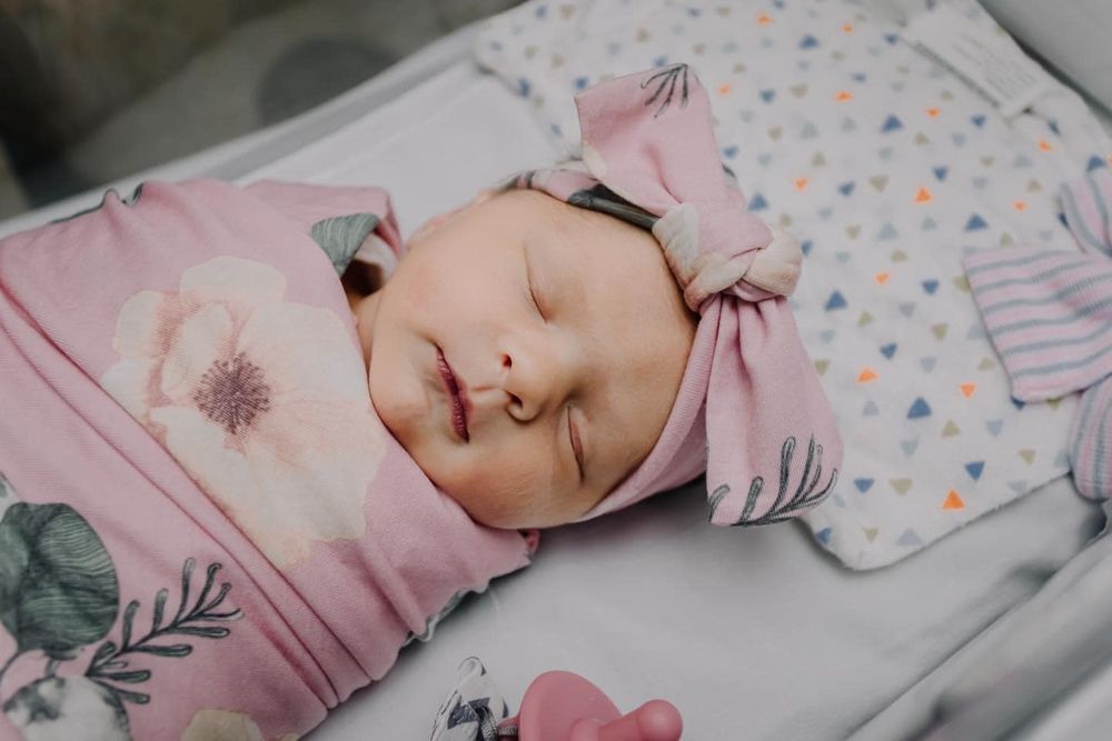 Peaceful sleeping newborn wearing pink floral swaddle and bow in hospital bassinet.