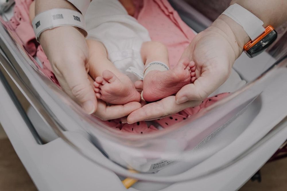 Hospital staff examines tiny newborn feet while baby rests on pink blanket.