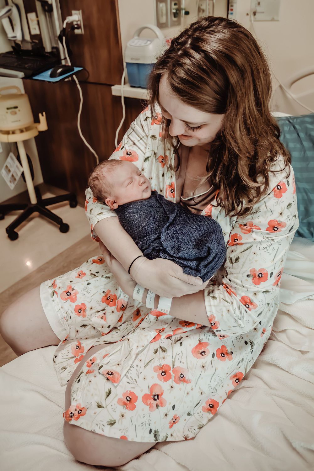 A woman in a floral dress holds and gazes lovingly at a newborn baby in a hospital room.