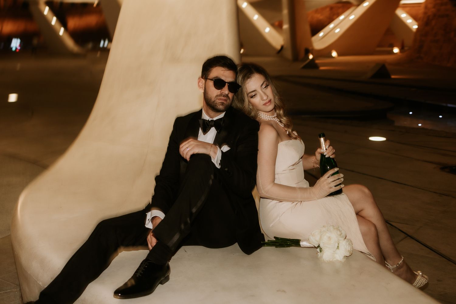 Couple in formal wear sits casually against curved wall with champagne.