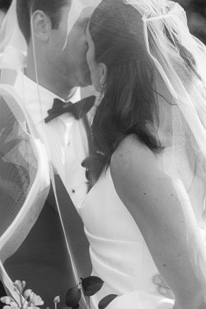 Black and white photo of a bride and groom sharing an intimate moment, veil draped over them, tuxedo and wedding dress.