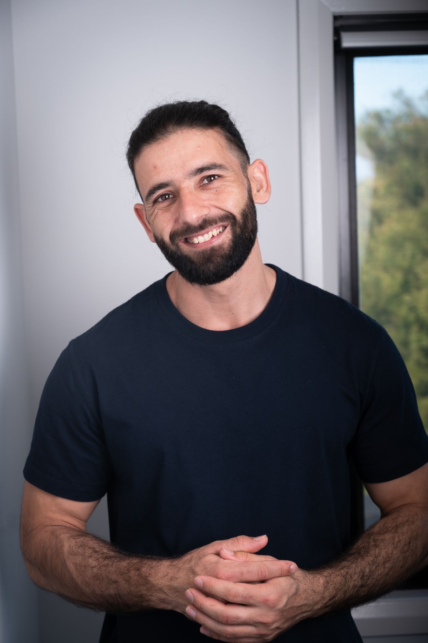 Smiling bearded man in a black t-shirt standing near a window with hands clasped.
